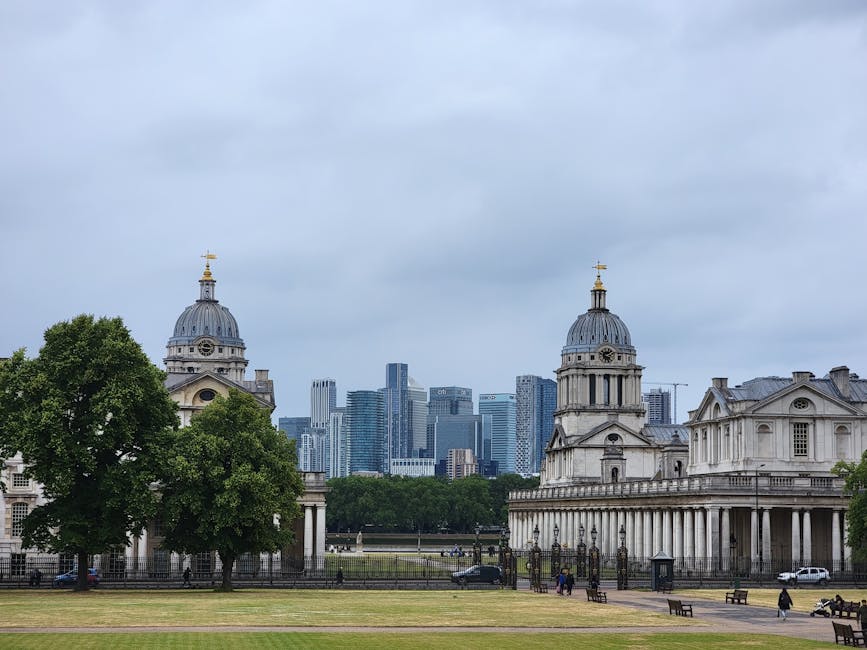 A wide view of historic buildings with classical architecture, including two prominent domed structures topped with gold finials, situated in a park with lush green trees and well-maintained grass. Modern high-rise office buildings and skyscrapers are visible in the background under an overcast sky, indicating a cityscape combining both historic and contemporary architecture. In the foreground, a paved area with benches and a few people walking or sitting is visible. The scene captures a typical city park environment near a central London relocation service, with elements suitable for home relocation or furniture transport, and highlights the process of moving within or out of Greenwich including loading and logistical planning at the site, consistent with services offered by Man With a Van Greenwich.