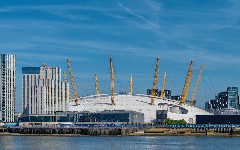 A large, modern sporting arena with a white, dome-shaped roof supported by multiple yellow pylons and cables, situated along the river Thames in Greenwich. Surrounding the arena are tall, contemporary office buildings with varied glass and steel facades. The scene is set during daytime with clear blue skies and a calm river in the foreground. The arena's exterior appears clean and well-maintained, indicative of a popular local landmark near Cutty Sark in Greenwich. This image captures the iconic structure as part of a cityscape, with potential relevance to home relocation or delivery services in the area, such as those provided by Man With a Van Greenwich, within the context of house removals and furniture transport.