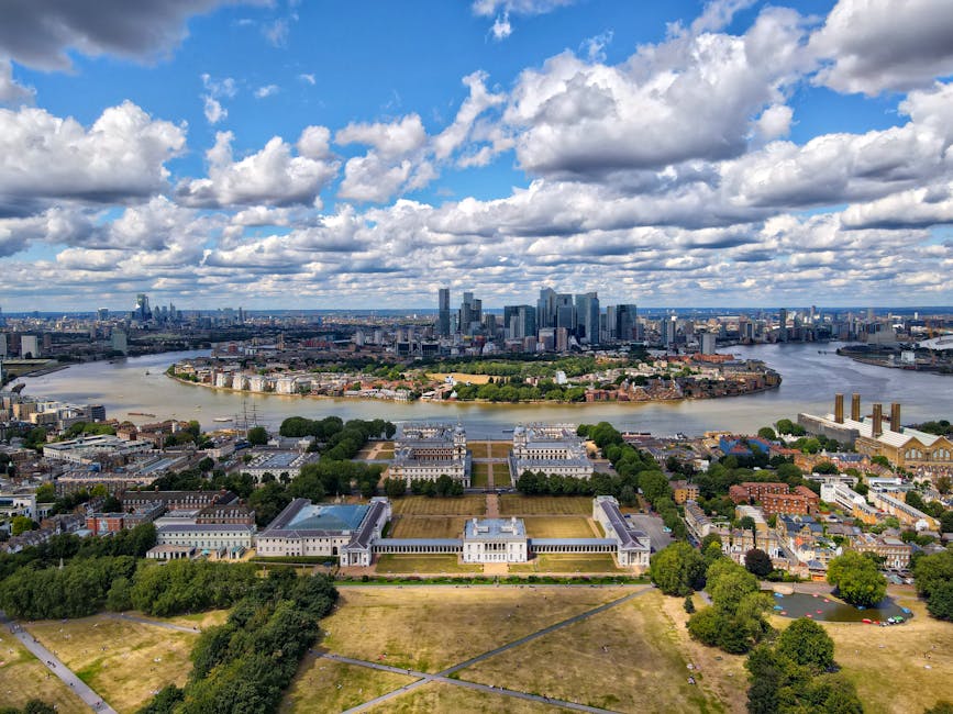 A wide view of historic buildings with classical architecture, including two prominent domed structures topped with gold finials, situated in a park with lush green trees and well-maintained grass. Modern high-rise office buildings and skyscrapers are visible in the background under an overcast sky, indicating a cityscape combining both historic and contemporary architecture. In the foreground, a paved area with benches and a few people walking or sitting is visible. The scene captures a typical city park environment near a central London relocation service, with elements suitable for home relocation or furniture transport, and highlights the process of moving within or out of Greenwich including loading and logistical planning at the site, consistent with services offered by Man With a Van Greenwich.