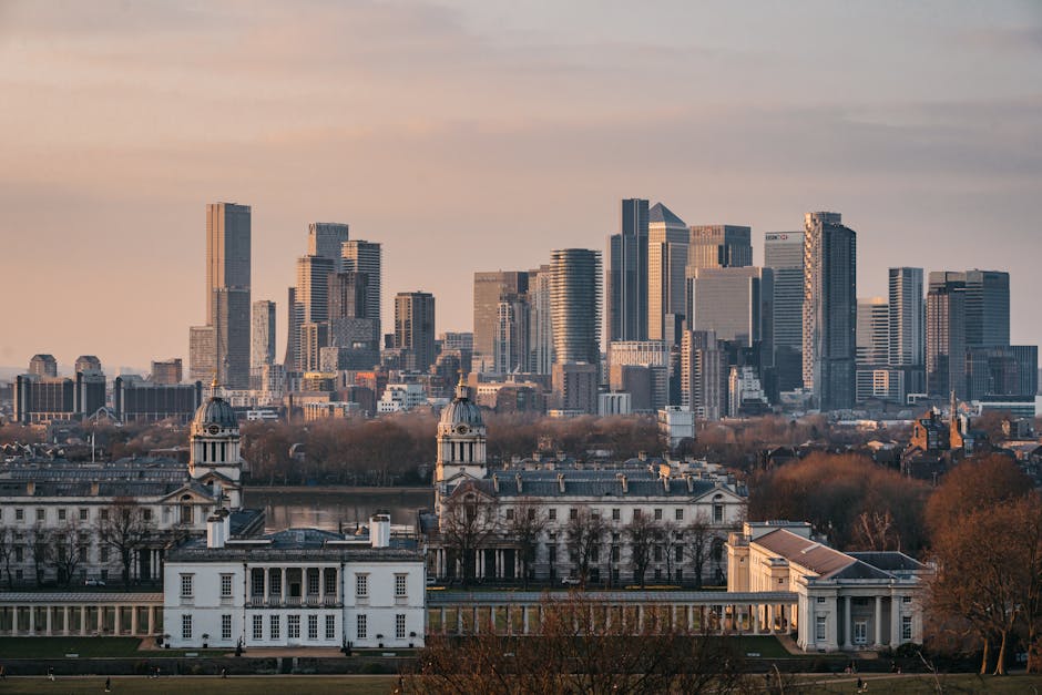 A panoramic view of London’s city skyline during late afternoon, showing tall modern skyscrapers and office buildings with glass facades reflecting the sunlight. In the foreground, there is a historic white building with classical architectural features, including columns and a dome, situated within a spacious park area with leafless trees and well-maintained lawns. The scene captures the contrast between the contemporary high-rise structures and the traditional architectural style of the historic building. This urban landscape illustrates the diverse architectural landscape typical of London, which is often involved in house removals and furniture transport services for home relocation projects, as evidenced by companies like Man With a Van Greenwich that specialise in such logistics within the city.