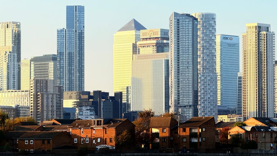 A cityscape featuring modern high-rise office buildings and residential towers with glass facades and sleek architecture, standing behind a row of traditional brick houses with pitched roofs and small front gardens. The buildings are bathed in natural daylight, highlighting the contrast between the urban skyscrapers and the older suburban homes. In the foreground, there are a few trees and parked cars along the street. This scene reflects a typical urban environment where house removals and home relocation services, such as those provided by Man With a Van Greenwich, facilitate the transport of furniture and belongings between residential areas and city centers, supporting seamless packing and loading processes.
