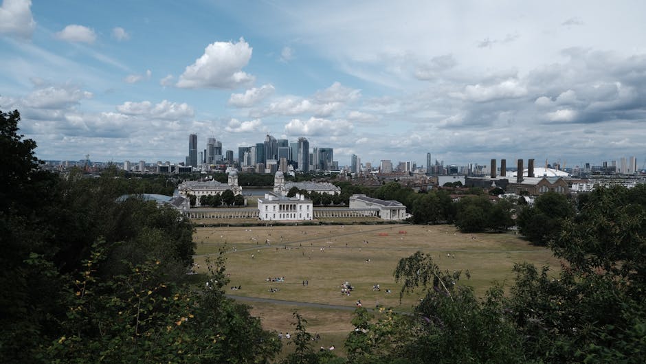 A wide view of Greenwich Park in London with several groups of people relaxing on the grassy open area, surrounded by trees and shrubbery in the foreground. In the distance, the historic Queen's House with its classical architecture is visible, set against the modern London skyline featuring tall skyscrapers and buildings. The sky overhead is partly cloudy, with a mix of white and grey clouds casting soft light over the scene. The image captures the outdoor environment during daylight hours, providing a natural setting for a home relocation or furniture transport process associated with house removals, with the city skyline in the background suggesting proximity to central London and supporting the context of moving services offered by Man With a Van Greenwich.