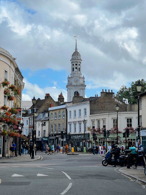 A busy street scene in Greenwich featuring a historic clock tower with a gold weather vane at the top, situated behind traditional brick and white-painted row of shops and buildings. The street is filled with pedestrians walking on the pavements and crossing at the intersection, some carrying shopping bags or bundled in jackets. On the right side, a delivery motorcycle and a car are parked near a lamp post. In the foreground, a loading area on the street is visible with a large wooden moving crate, cardboard boxes wrapped in plastic, and furniture covered with protective blankets, indicating preparations for a home relocation. A man from Man With a Van Greenwich is seen lifting or positioning packing materials as part of the furniture transport process, while other personnel are organizing items for efficient loading into a van parked parallel to the pavement. The sky above is partly cloudy, with natural daylight illuminating the scene, reflecting a typical day of packing and moving activities in the area.