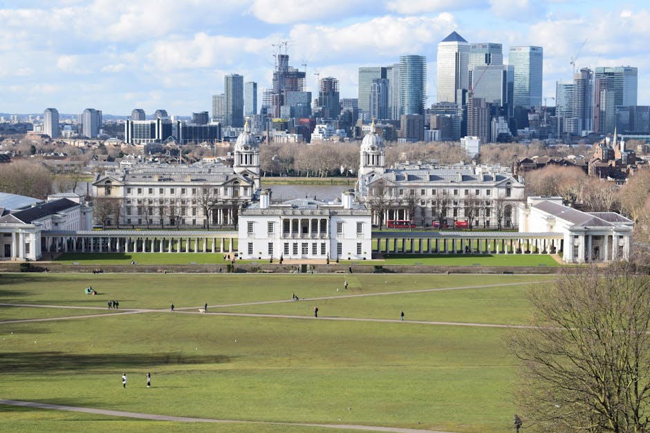 A wide view of Greenwich Park in London with several groups of people relaxing on the grassy open area, surrounded by trees and shrubbery in the foreground. In the distance, the historic Queen's House with its classical architecture is visible, set against the modern London skyline featuring tall skyscrapers and buildings. The sky overhead is partly cloudy, with a mix of white and grey clouds casting soft light over the scene. The image captures the outdoor environment during daylight hours, providing a natural setting for a home relocation or furniture transport process associated with house removals, with the city skyline in the background suggesting proximity to central London and supporting the context of moving services offered by Man With a Van Greenwich.
