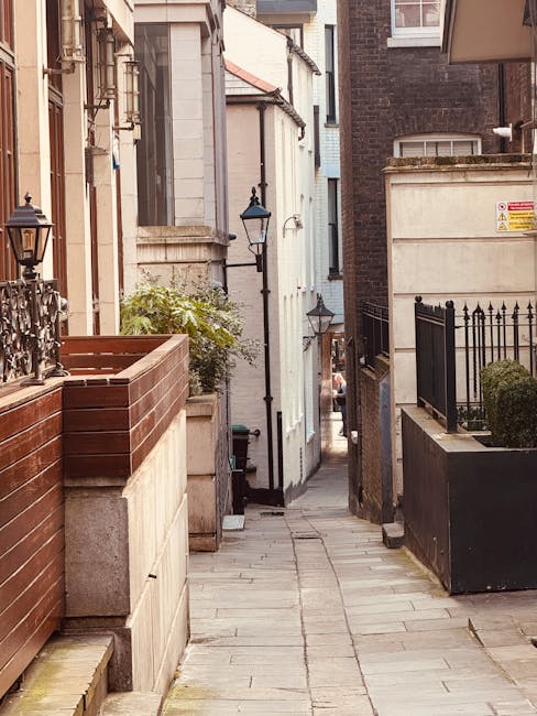 A narrow residential alleyway with a paved stone path between closely situated buildings featuring white and dark brick exteriors. On the left, there is a wooden planter box with a small green shrub and a vintage-style black lamp post attached to the wall. On the right, a black metal fence with decorative vertical bars encloses a small garden area with a neatly trimmed shrub. The alleyway shows a slight downward slope, with a vanishing point towards the background where more buildings are visible. This setting illustrates the typical access challenges faced during house removal or furniture transport on tight streets in Greenwich, and is relevant to the services offered by Man With a Van Greenwich, highlighting the importance of careful loading and unloading procedures in constrained urban environments.