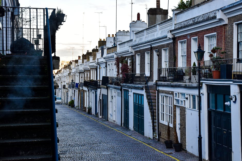 A narrow residential alleyway with a paved stone path between closely situated buildings featuring white and dark brick exteriors. On the left, there is a wooden planter box with a small green shrub and a vintage-style black lamp post attached to the wall. On the right, a black metal fence with decorative vertical bars encloses a small garden area with a neatly trimmed shrub. The alleyway shows a slight downward slope, with a vanishing point towards the background where more buildings are visible. This setting illustrates the typical access challenges faced during house removal or furniture transport on tight streets in Greenwich, and is relevant to the services offered by Man With a Van Greenwich, highlighting the importance of careful loading and unloading procedures in constrained urban environments.