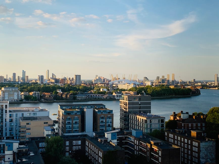 A wide-angle view of a cityscape taken from an elevated perspective during daylight, showcasing a mix of modern high-rise buildings and older low-rise structures along a river. In the foreground, there are several residential buildings with varying architectural styles, including balconies and flat roofs, some of which are covered with solar panels and rooftop equipment. The middle ground features a calm river with a few small boats visible, separating the residential area from the distant skyline. Across the river, a cluster of taller office towers and commercial buildings can be seen, with some under construction, evidenced by cranes. The sky is mostly clear with a few wispy clouds, and the warm sunlight highlights the various building details and river reflections. This scene relates to home and office relocations with potential views of nearby transportation hubs and urban infrastructure, relevant to [COMPANY_NAME]'s house removals and furniture transport services in the Greenwich area.