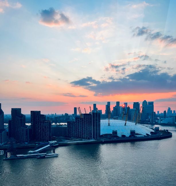 A panoramic view of the North Greenwich area during sunset, showcasing the distinctive O2 Arena with its white, tent-like roof structure situated by the river Thames. To the left, there are several modern high-rise buildings made of dark glass and steel, reflecting the fading sunlight. The sky is painted with soft pastel hues of pink, orange, and blue, with scattered clouds overhead. The river below is calm, with a small dock and boats visible along the waterfront. In the foreground, the edge of a property’s interior is partially visible, with a glimpse of a blue wall. This scene captures the urban landscape relevant to a house removals case study conducted by Man With a Van Greenwich, focusing on local transport and relocation logistics in Greenwich during an office move to North Greenwich O2, with the city skyline providing context for the moving process.