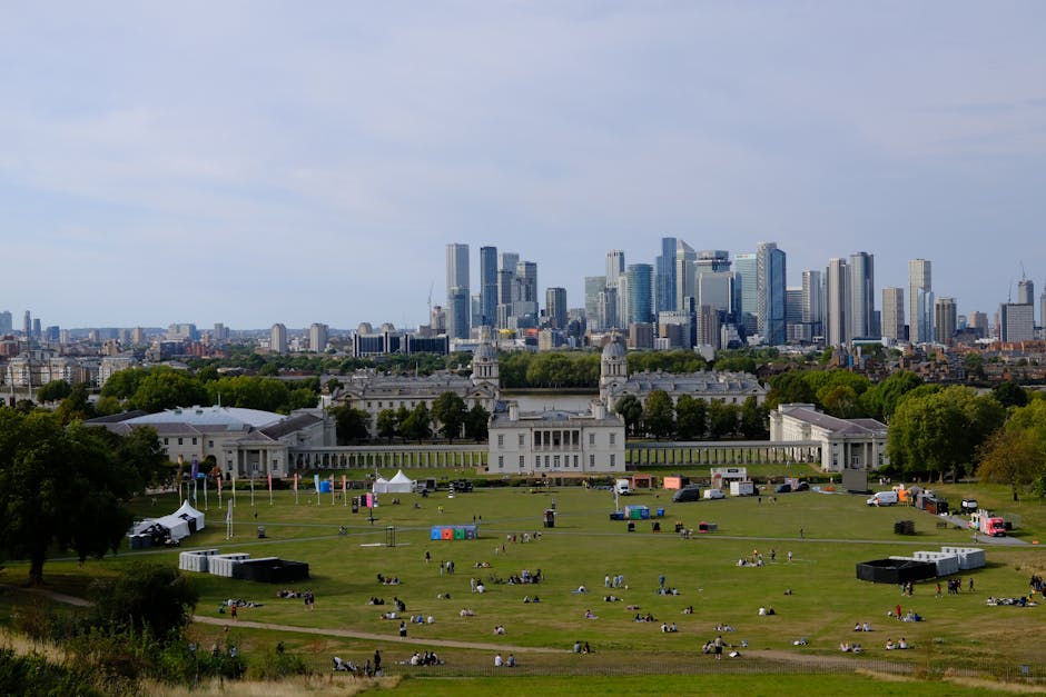 A wide view of Greenwich Park in London on a clear day, with lush green open space populated by groups of people sitting and relaxing on the grass, some near picnic setups. In the middle ground, the historic Queen's House with its white classical facade and twin clock towers is visible, surrounded by trees and pathways. In the background, the modern London skyline features tall glass skyscrapers and office buildings, creating a contrast between historic and contemporary architecture. The scene includes vehicles parked along the park’s edge, and there are small tents and equipment set up, suggesting preparations for outdoor events. The lighting is bright with a partly cloudy sky, highlighting the different elements of nature, architecture, and urban development. This image could be used to illustrate the logistical process of home relocation and furniture transport, emphasizing the importance of planning and executing moving services efficiently, such as those offered by Man With a Van Greenwich, especially for urgent moves like same-day removals in Greenwich.